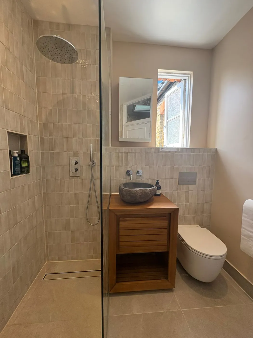 Walk-in shower with rainfall head, tiled niche, teak vanity with stone countertop basin and wall-hung WC in a beige-tiled family bathroom.