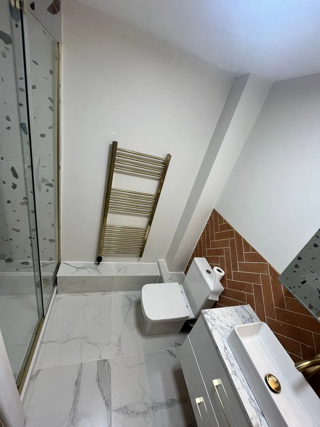 En-suite shower room with terracotta herringbone feature tiles, marble floor, gold tap and white countertop basin.