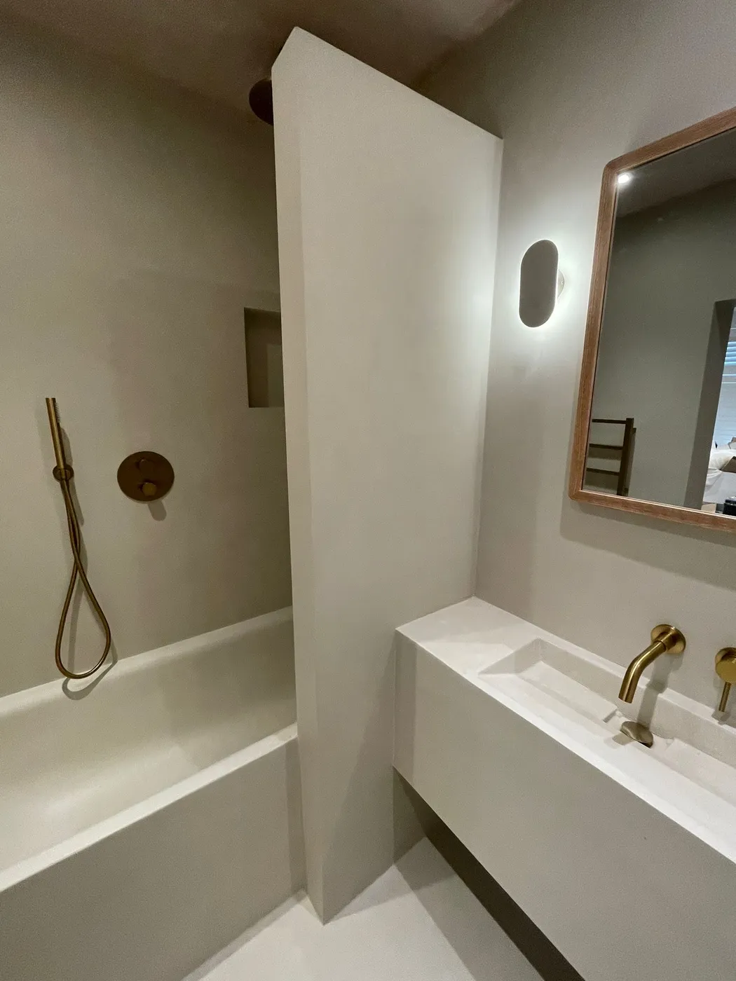 Seamless microcement bathroom in warm taupe with brushed brass shower fittings, integrated basin ledge and wood-framed mirror.