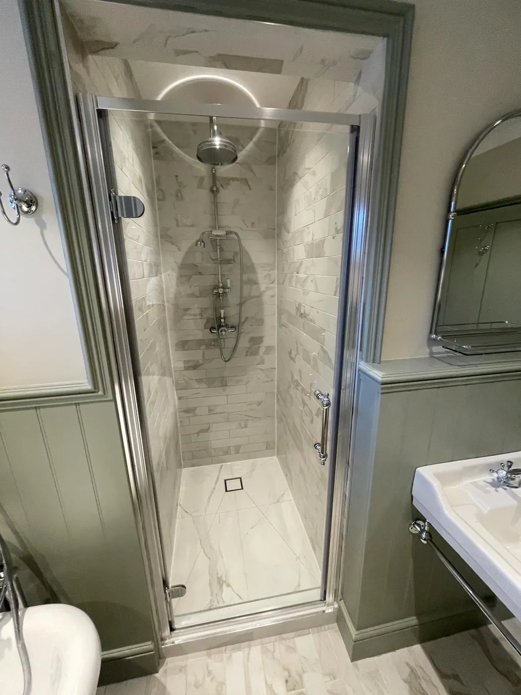 Traditional shower room with sage green wall panelling, marble-effect tiled shower cubicle, arched mirror and pedestal basin.