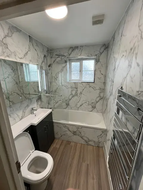 Family bathroom renovation with marble-effect wall panels, fitted bath with shower over, dark vanity unit and wood-effect floor.