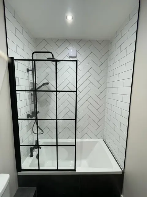 Over-bath shower with black Crittall-style grid screen, matt black fittings and white herringbone tiles in a modern London bathroom.